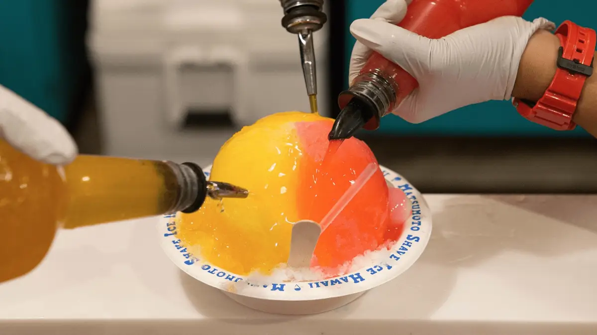 A close-up of hands wearing gloves pouring vibrant yellow, red, and orange syrups from squeeze bottles onto a large mound of shaved ice in a bowl—a delicious representation of foods that define Hawaiian culture. A wooden spoon rests on the side, and the text on the bowl's rim says "HOT SPOON ICE HAWAII.