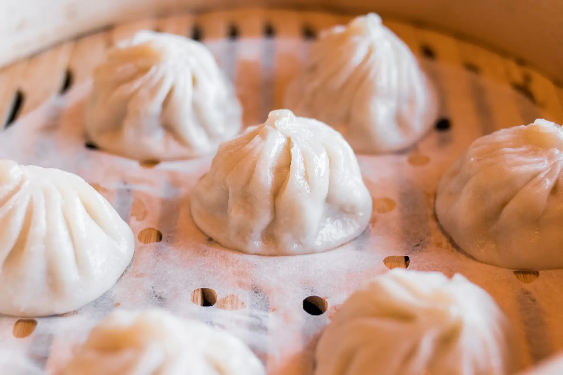 Close-up image of several steamed dumplings resting on a parchment-lined bamboo steamer. These delectable bites, emblematic of foods that define Hawaiian culture with international influences, are round with pinched tops, showcasing their delicate and soft dough. The steamer's small holes ensure even cooking.