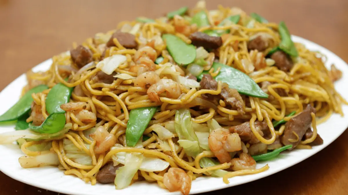 A plate of chow mein, rich in international influences, is topped with shrimp, sliced beef, cabbage, and snow peas. The dish features stir-fried noodles mixed with the various ingredients, creating a colorful and appetizing presentation. The background is blurred, focusing attention on the plate.