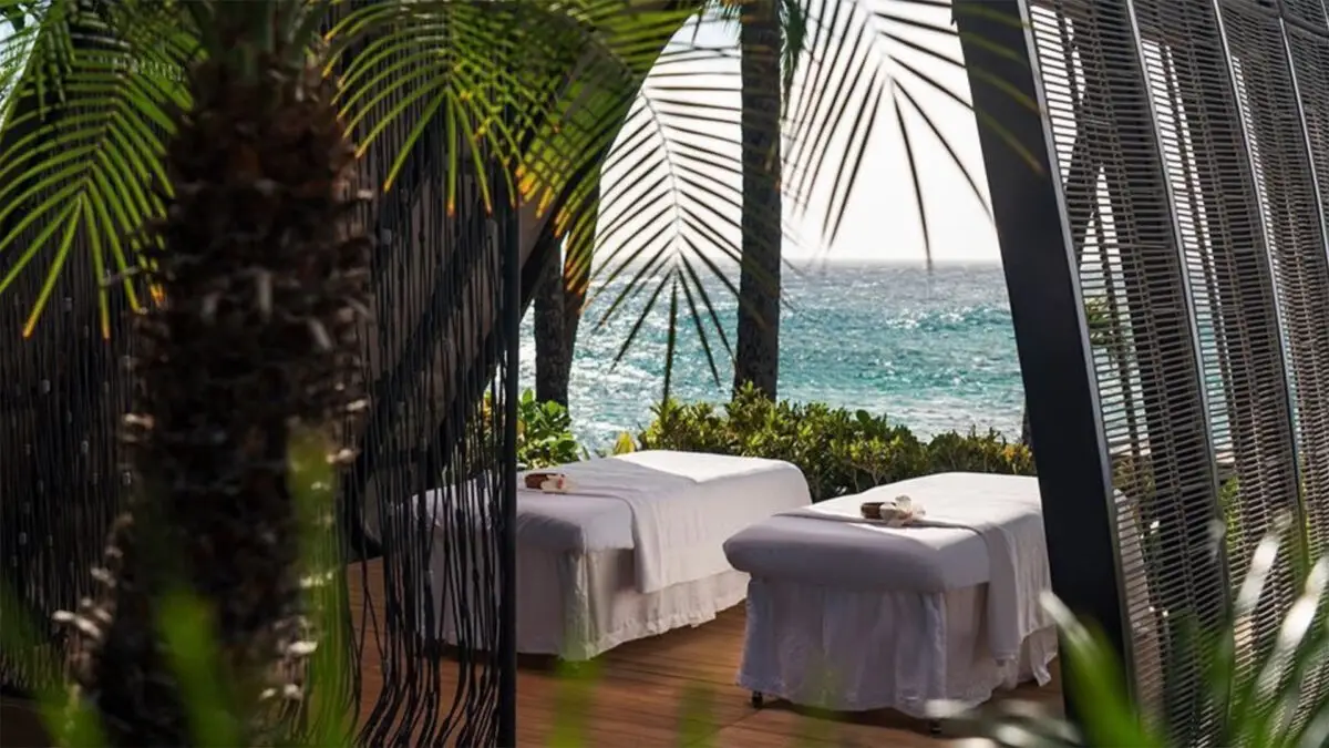 Two massage tables covered with white sheets are situated under a canopy by the sea, surrounded by lush greenery and palm fronds. The ocean waves and blue sky create a serene and relaxing atmosphere, epitomizing the experience offered by the best spas Maui has to offer.
