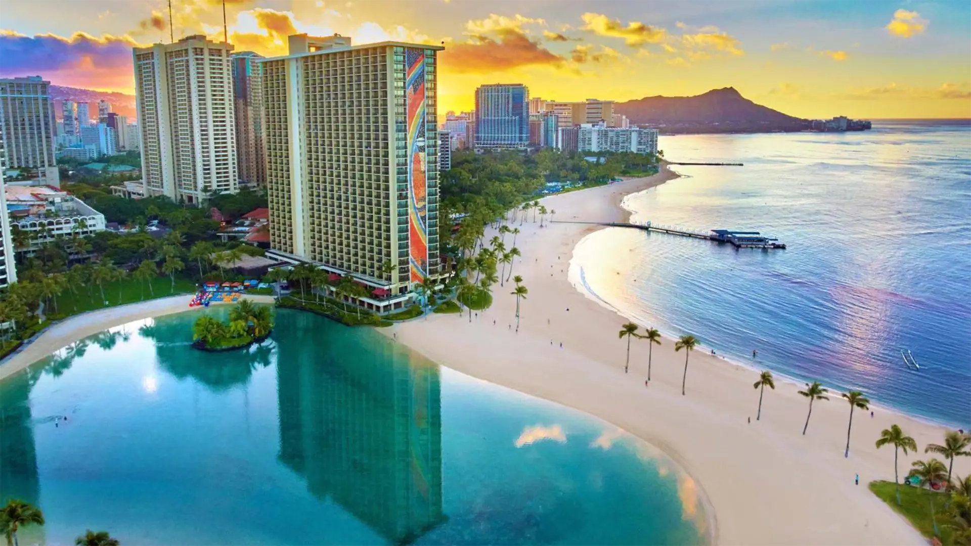 Aerial view of a picturesque beachside city at sunset. High-rise buildings including some of the best hotels on Oahu line the coast, with a curving sandy beach and calm turquoise waters in front. A mix of palm trees and greenery is scattered around, with a mountain visible in the distance.