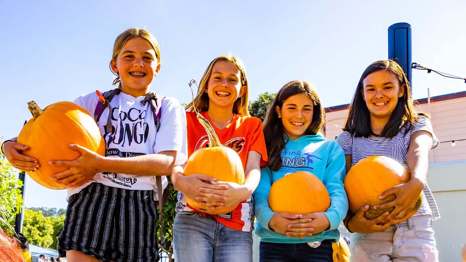 Four smiling kids stand in a row under a clear sky, each holding a pumpkin. They appear happy and excited, dressed in casual clothes, enjoying a bright, sunny day—truly one of the best things to do in South Bay this October.