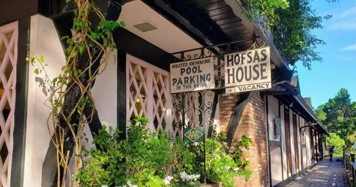 A quaint, rustic inn named "Hofsas House" with a vacancy sign. The exterior is adorned with climbing plants and flower boxes, featuring wooden beams and lattice windows. Signs for amenities like a heated swimming pool and parking are visible. Ideal for those seeking romantic hotels in Carmel, it is surrounded by trees and greenery.