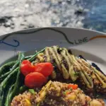 A close-up of the best dinner in Kona by the sea. The dish features grilled fish topped with a cream sauce, green beans, red cherry tomatoes, and a serving of colorful quinoa. The plate is white with a decorative design, and sunlight reflects off the water in the background.