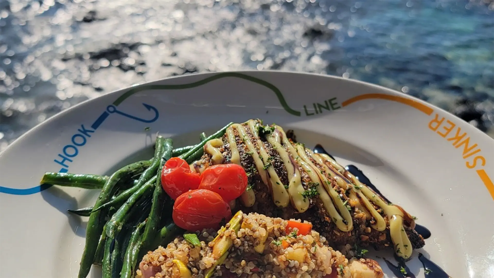 A close-up of the best dinner in Kona by the sea. The dish features grilled fish topped with a cream sauce, green beans, red cherry tomatoes, and a serving of colorful quinoa. The plate is white with a decorative design, and sunlight reflects off the water in the background.