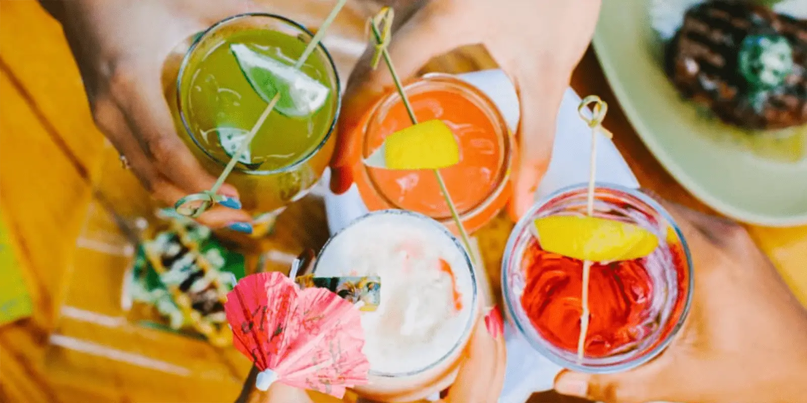 A top-view photo showing four hands holding colorful drinks in glasses, garnished with various decorations like umbrellas, lemon wedges, and fruit slices. The background has blurred food items on a wooden table, capturing the best Happy Hour vibes in Kaanapali Maui.
