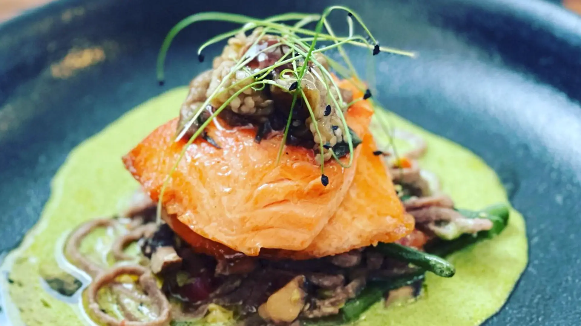 A close-up of an elegantly plated gourmet dish featuring a beautifully seared salmon fillet atop a bed of sautéed mushrooms, green beans, and soba noodles, garnished with a medley of microgreens. Enjoy the best dinner in Lihue with this delicacy surrounded by a vibrant green sauce.