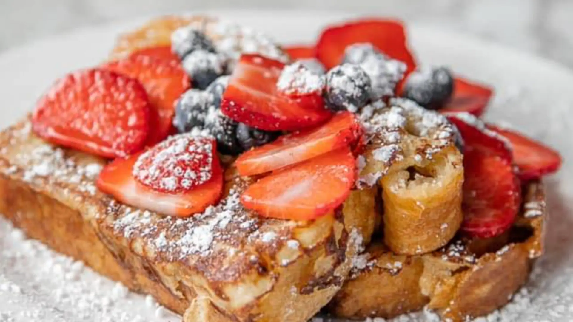 A plate with French toast topped with powdered sugar, sliced strawberries, and blueberries. The French toast is golden brown and looks fluffy and appetizing, with the fresh berries and powdered sugar adding vibrant color and an element of sweetness - truly the best breakfast in Lihue.