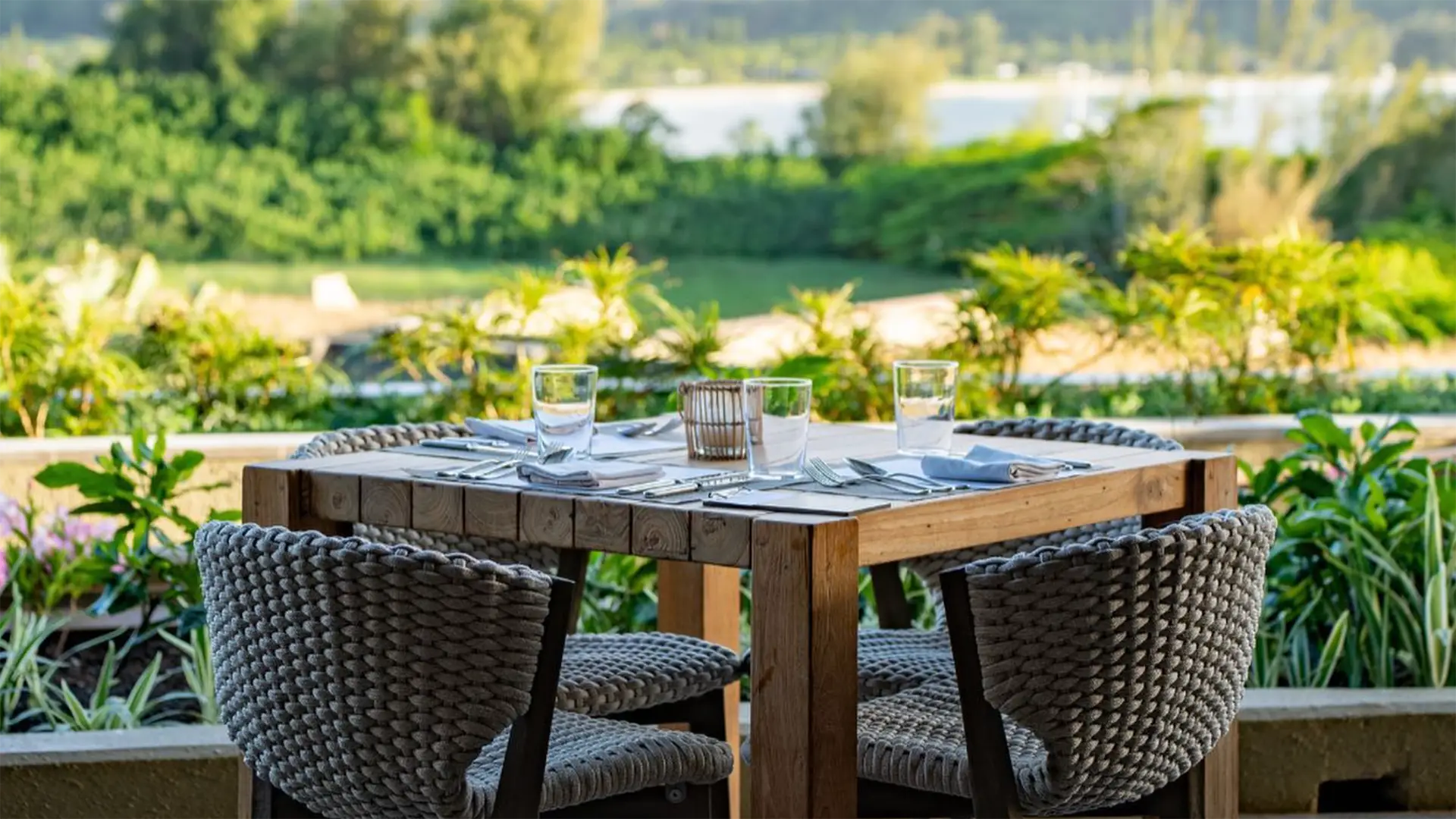 An outdoor table set for four, with glasses and napkins, surrounded by woven chairs. The background features lush greenery, a river, and hills under a clear sky, creating a serene setting perfect for enjoying the best breakfast in Kauai.