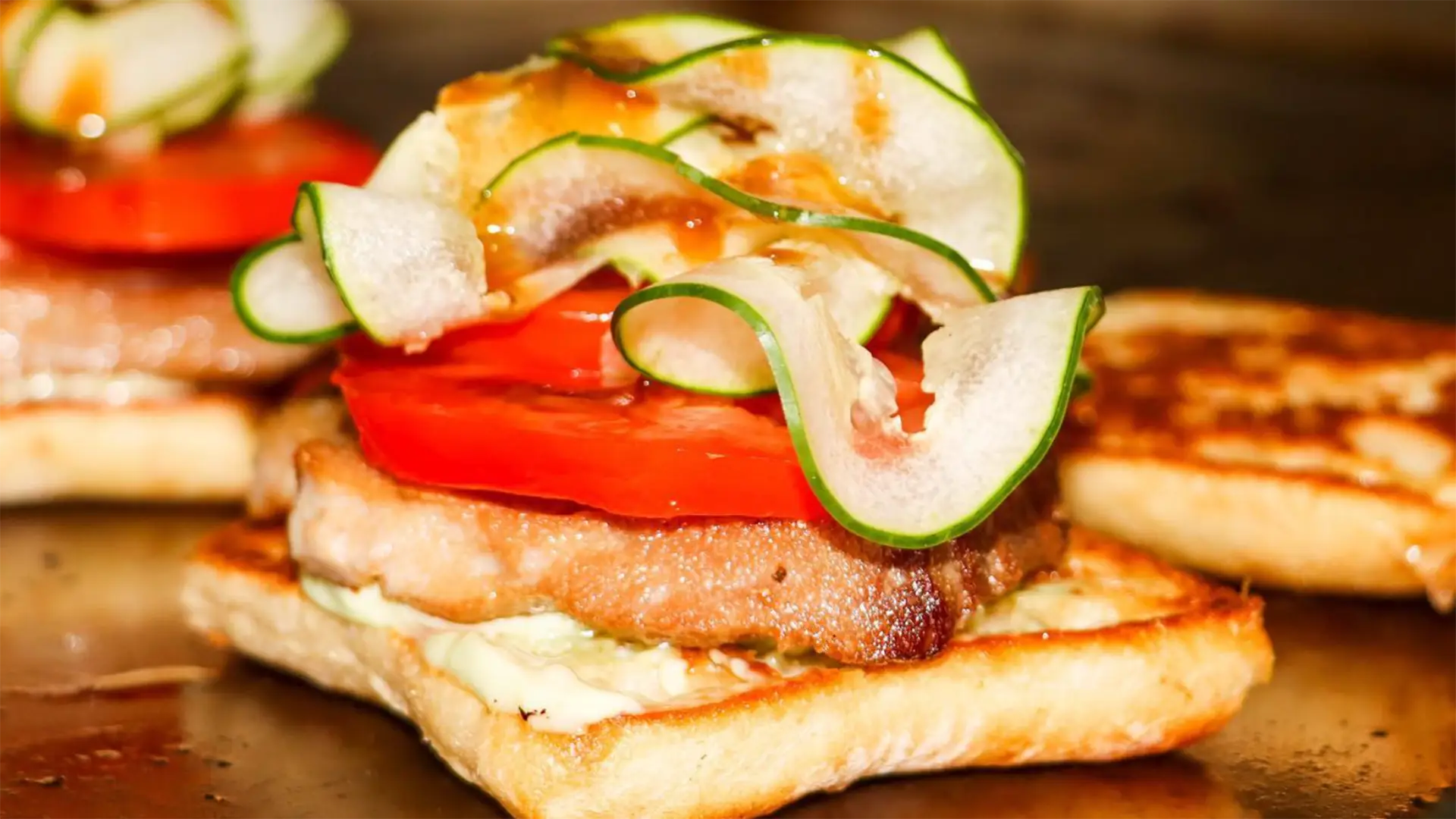 A close-up of an open sandwich, perfect for the best breakfast on Kauai's South Shore, featuring a thick slice of grilled meat, fresh tomato slices, and thinly sliced cucumber and onion atop a slice of toasted bread. The sandwich is topped with a drizzle of sauce.
