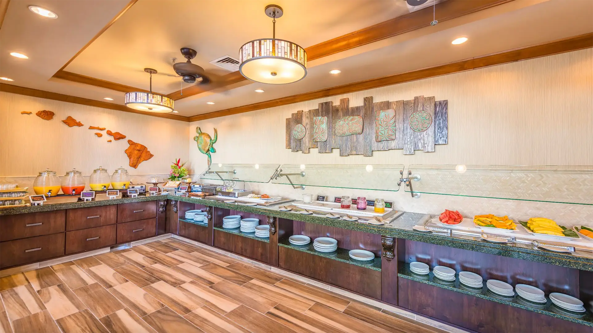 A well-lit buffet with a variety of dishes on display, including fruits, desserts, and condiments. The room has wooden floors and warm lighting. A wall features wooden carvings, tropical decor, and overhead lights, creating an inviting atmosphere. Enjoy the best breakfast Kauai South Shore offers here!