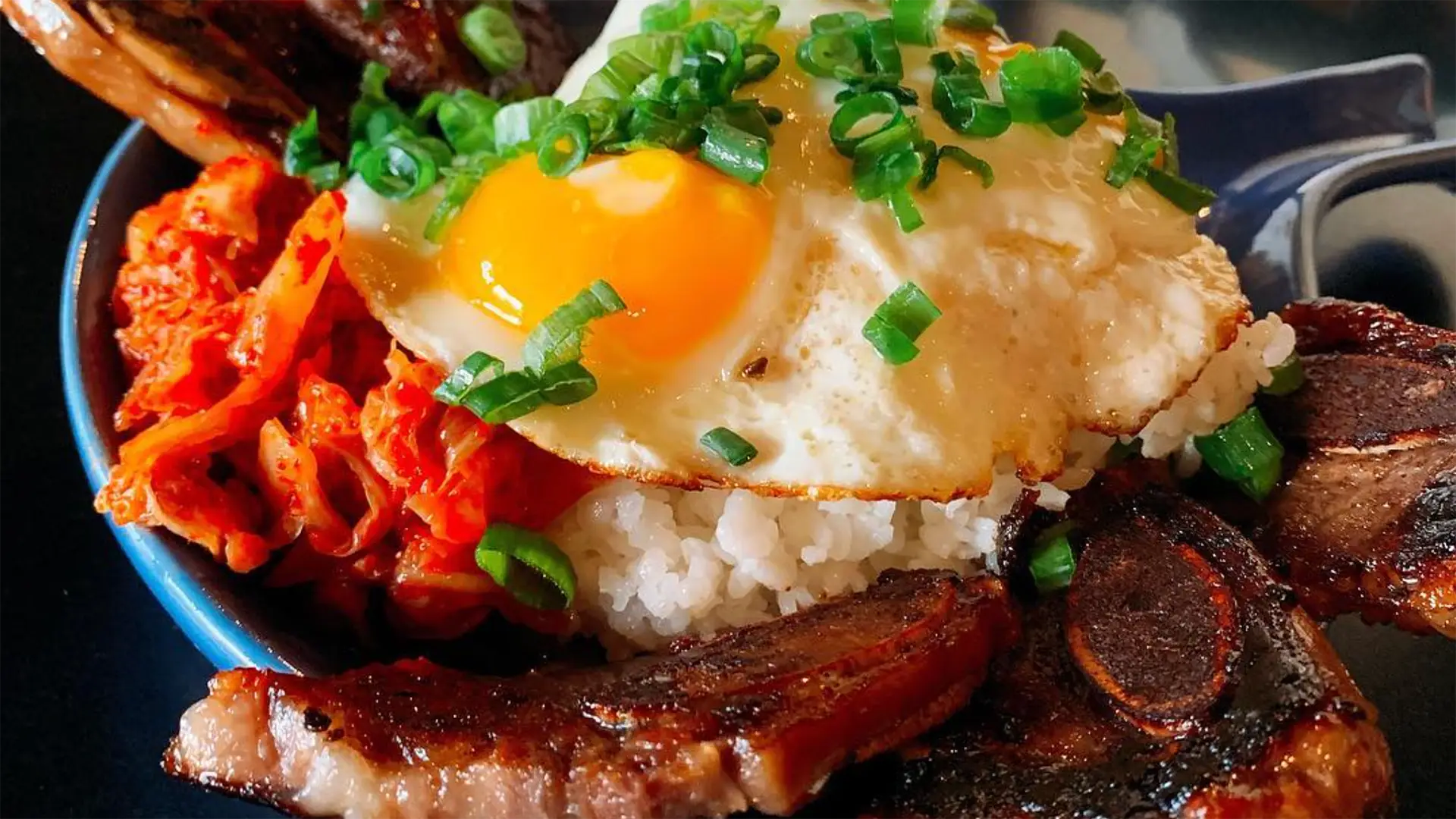 A bowl filled with white rice, topped with a sunny side up egg garnished with chopped green onions, slices of grilled meat, and a portion of kimchi on the side. The vibrant dish boasts rich, appetizing colors and textures—truly one of the best breakfast finds in Kauai.