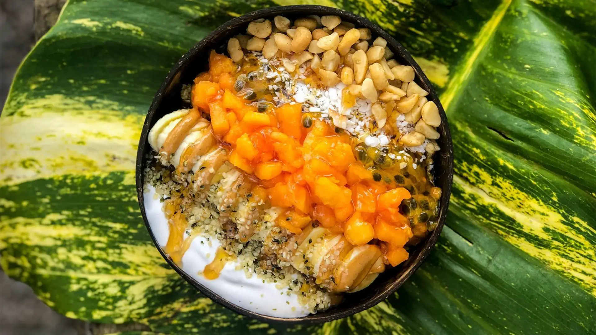 A vibrant smoothie bowl on a green, leafy background, topped with chunks of fresh mango, passionfruit, banana slices, chopped nuts, shredded coconut, and hemp seeds, creating a colorful, appetizing display.