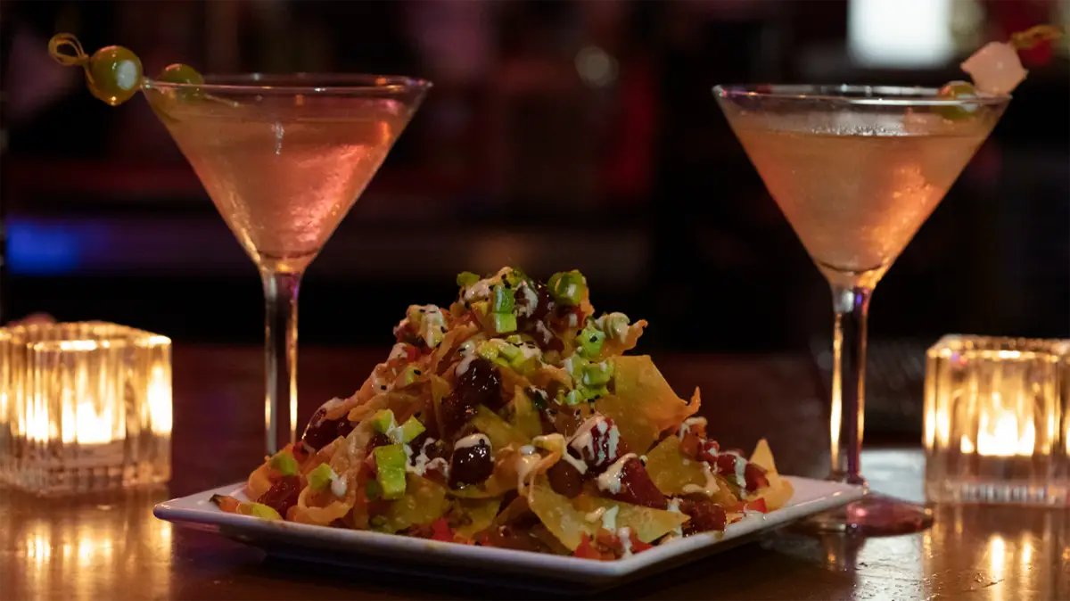 A plate of loaded nachos topped with green onions, cheese, and sauce is on a wooden table. Two martini glasses with light-colored cocktails, each garnished with olives, are positioned on either side of the plate. Dim, ambient lighting in the background sets the perfect mood for Kauai's best happy hour on the Central Coast.