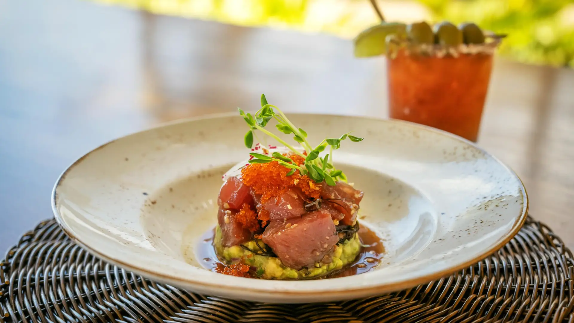 A gourmet dish featuring diced raw tuna on a bed of avocado, garnished with microgreens and roe, presented on a white plate with a decorative rim. In the background, there is a drink with a skewer of olives and a lime slice—perfect for enjoying the best happy hour Kauai has to offer.