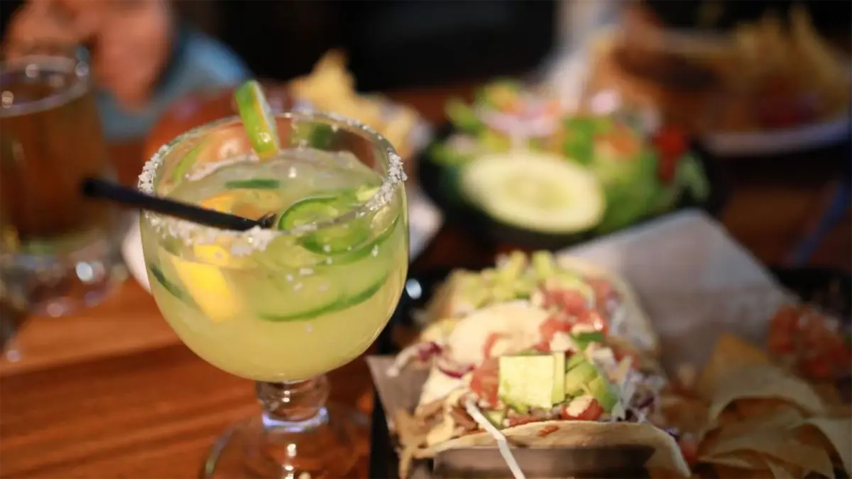 A close-up of a table setting with a margarita garnished with lime slices and a salted rim in focus. The background features a blurred view of tacos filled with various toppings and a salad, suggesting the best happy hour in Kauai for enjoying a Mexican meal.