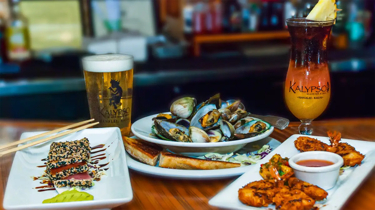 A spread of seafood dishes on a wooden table, including a plate of seared sesame tuna, a bowl of steamed clams, fried shrimp with dipping sauce, and drinks like beer and a pineapple-garnished tropical cocktail. The setting appears to be a restaurant offering the best happy hour on Kauai's North Shore.