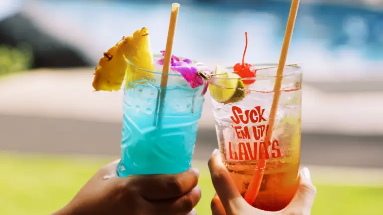 Close-up of two hands holding colorful tropical drinks in clear plastic cups with straws. The left drink is blue with a pineapple slice and pink flower garnish, while the right drink is orange-red with a cherry and lime garnish. A pool is visible in the background—best happy hour on Kauai's South Shore.