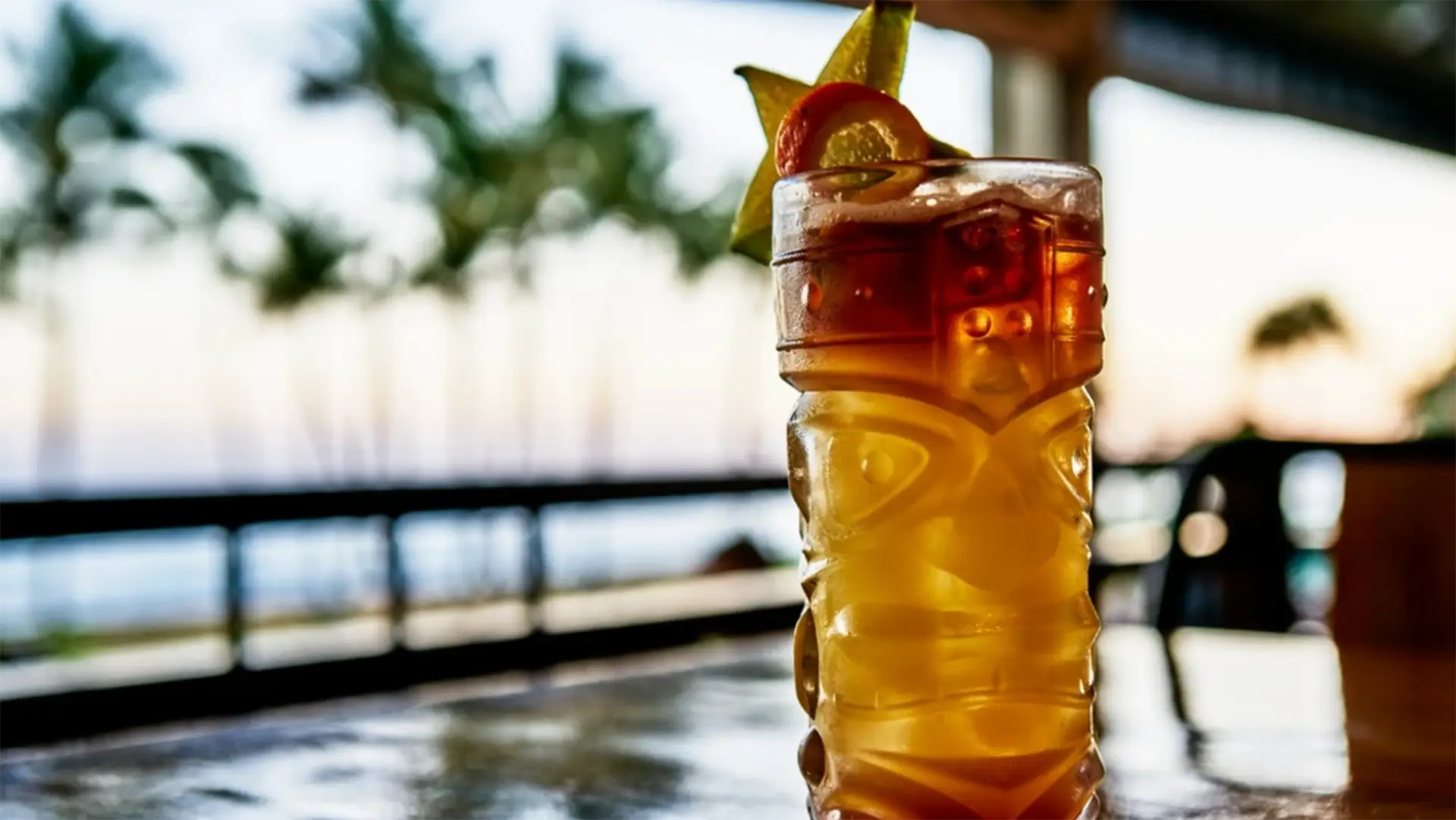 A tropical cocktail in a tall patterned glass is garnished with a slice of lime and a cherry. The drink is placed on a shiny surface with a background showing blurred palm trees and an outdoor setting.