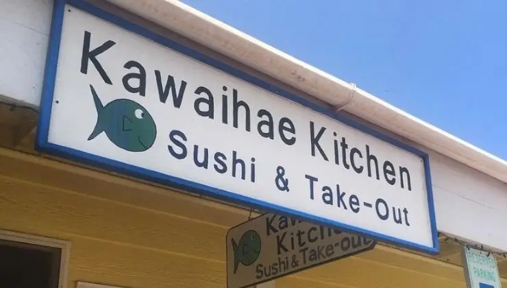 Sign for "Kawaihae Kitchen" with a fish logo, and the text "Sushi & Take-Out" beneath, renowned for having the best sushi on the Big Island. The sign is mounted on a building with light yellow walls, and there's a clear blue sky in the background.