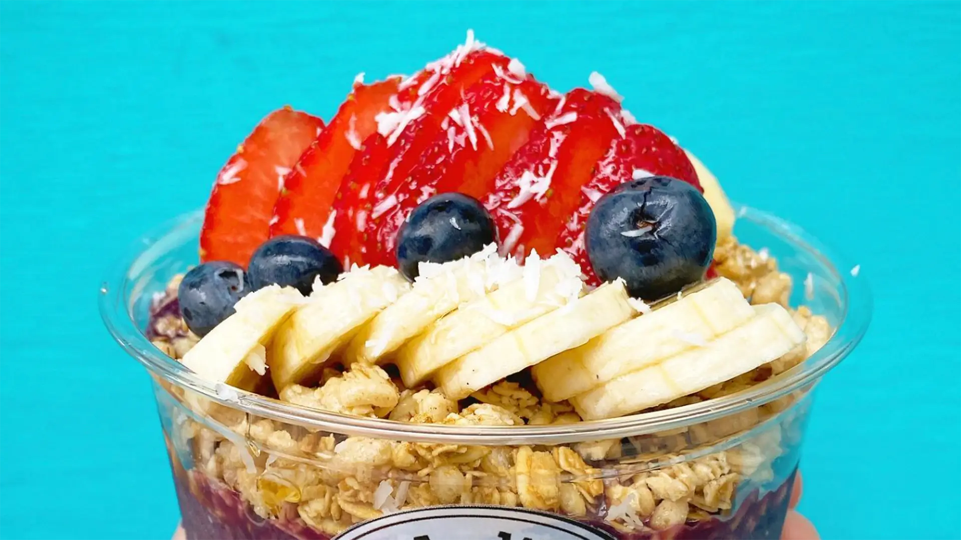 A colorful acai bowl topped with fresh fruit, reminiscent of the best smoothies on the Kona Big Island, Hawaii. Sliced strawberries and bananas are arranged neatly beside whole blueberries. The bowl also contains granola and a sprinkle of shredded coconut, all set against a vibrant turquoise background.