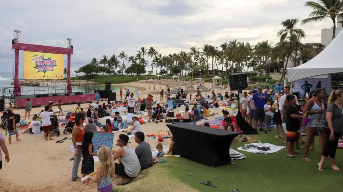 A large crowd gathers on a beach and grassy area, some seated on towels and blankets, enjoying an outdoor event. A stage with a banner reading "Island Music Festival" is set up in the background. Palm trees line the edge of the beach under a partly cloudy sky, celebrating one of Ko Olina's Annual Events.