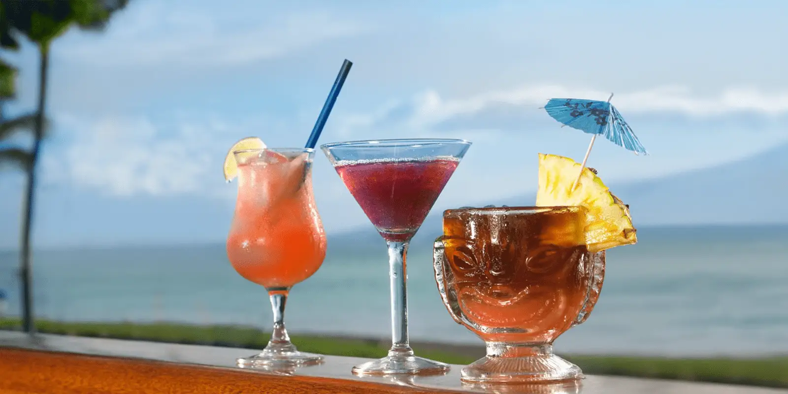 Three tropical cocktails grace a wooden surface with Kaanapali maui's beach and ocean in the background. The best Happy Hour features a pink cocktail with a lemon slice and straw, a martini glass holding a purple drink, and a brown tiki glass adorned with a pineapple slice and mini umbrella.