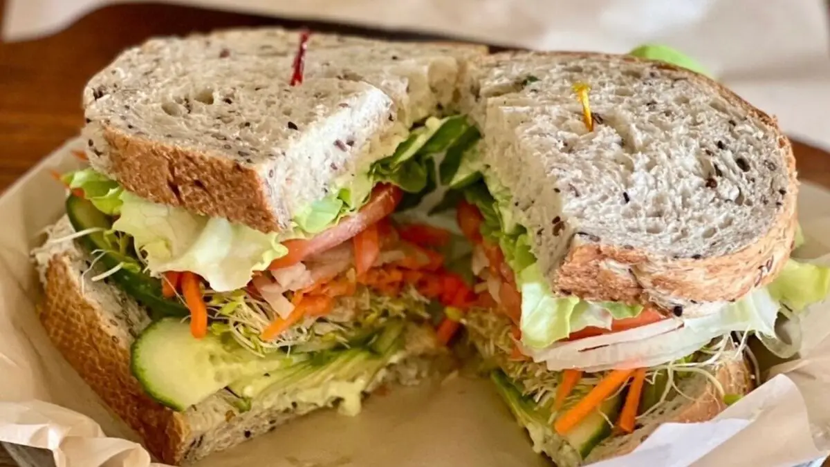 A Maui-inspired sandwich made with seeded whole-grain bread, filled with fresh vegetables including lettuce, tomato, cucumber, carrots, and sprouts. It's cut in half and held together with toothpicks, then placed on parchment paper.