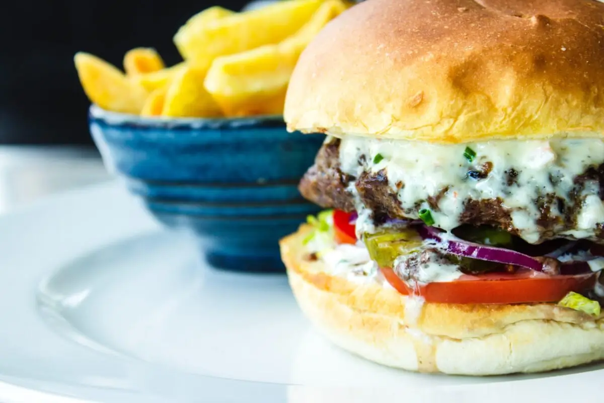 A close-up of a juicy burger with melted cheese, lettuce, tomato, onions, and pickles in a soft bun on a white plate. This mouthwatering creation hails from one of the best burger spots in Lihue. In the background, there is a side of golden, crispy fries in a blue bowl.