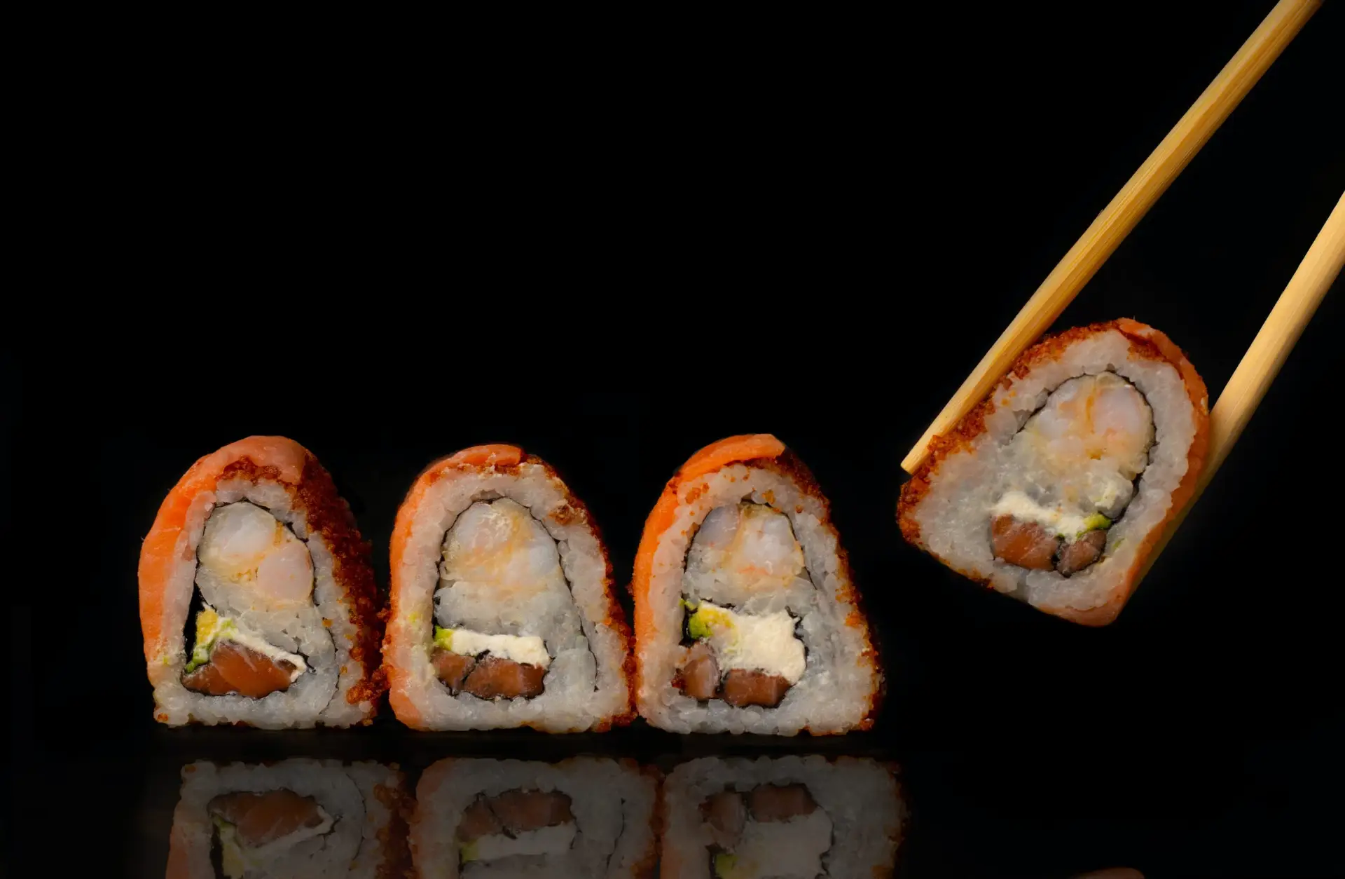 Four pieces of sushi roll with a crunchy coating and a variety of fillings are lined up on a reflective surface against a black background. The piece on the right is being held by a pair of chopsticks, showcasing what might be the best sushi in Poipu or Koloa.
