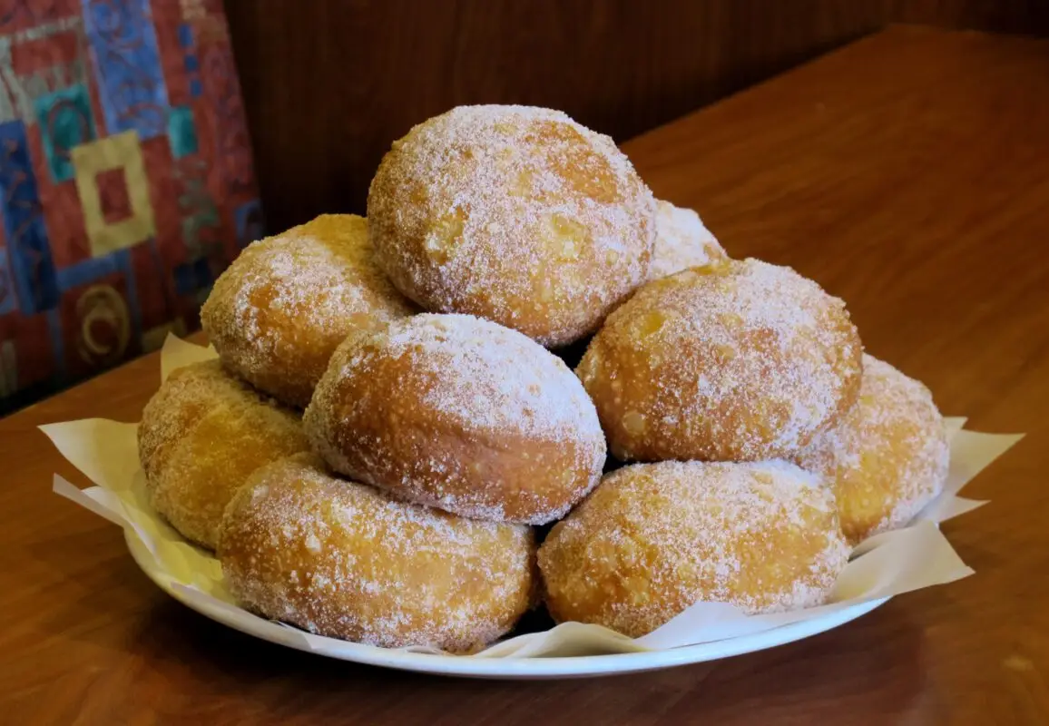 A plate filled with several sugar-coated doughnuts is placed on a wooden table. The doughnuts are stacked into a small mound, creating an appealing display of freshly baked treats.