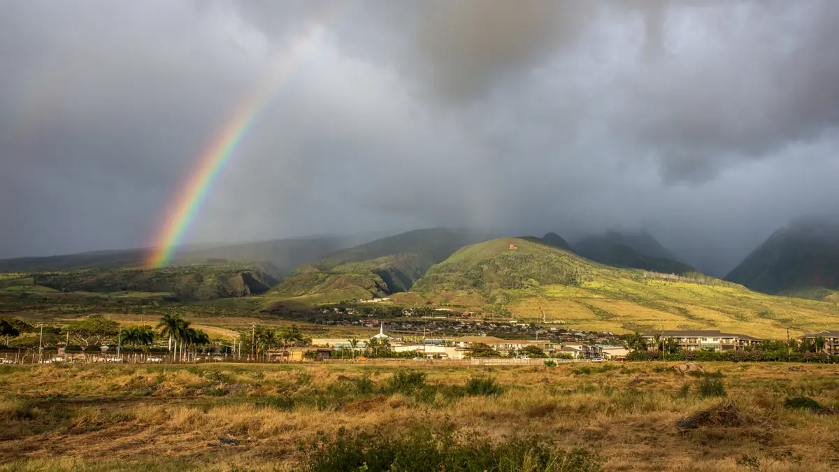 A scenic landscape depicts a vibrant rainbow arching across a cloudy sky over lush green hills. The scene evokes ideas of what to do on Maui when it rains, with the small town nestled among greenery and open fields contrasting dramatically with the sky and majestic mountains in the background.