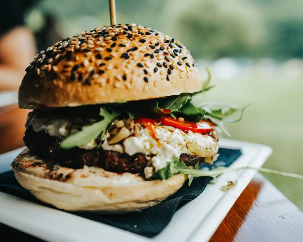 A close-up of one of the best burgers in Poipu, with a seeded bun, filled with a beef patty, arugula, tomatoes, feta cheese, and other vegetables. Served on a white plate with a wooden skewer holding it together. The background is blurred.