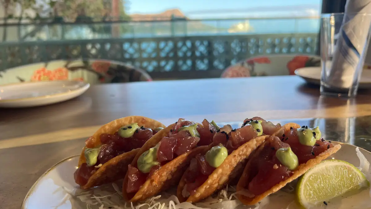A plate of four ahi poke tacos garnished with green sauce and sesame seeds sits on a table. A lime wedge accompanies the tacos. In the background, Oahu's best sunset dining offers an outdoor seating area with a breathtaking view of the ocean and distant mountains.