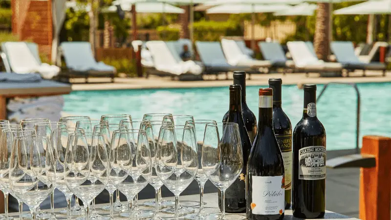 A poolside scene with several rows of empty wine glasses on a table, along with several bottles of red and white wine, evokes the best things to do in Napa Valley November. In the background, lounge chairs with umbrellas are arranged around a sparkling blue pool.