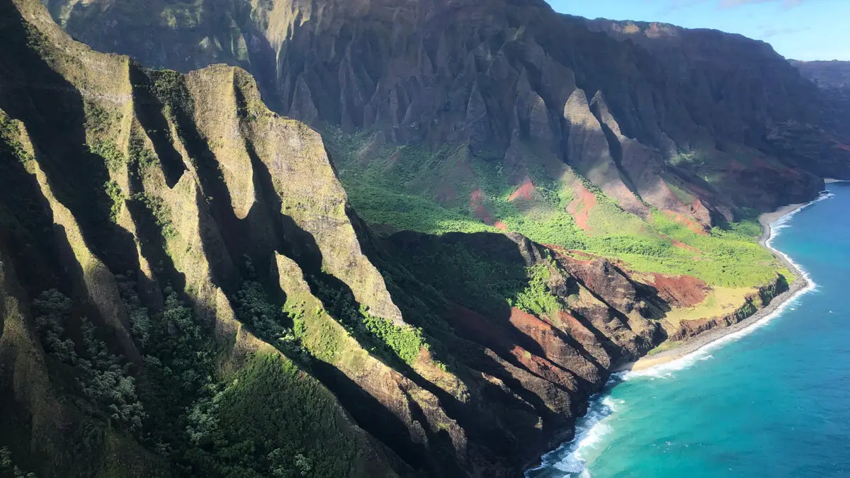 Aerial view of the Na Pali Coast on the island of Kauai, Hawaii. As one of the best places to visit on Kauai, the rugged coastline features dramatic cliffs, deep green valleys, and a turquoise ocean with white waves crashing on the shore. Sunlight casts shadows on the steep, jagged mountains.