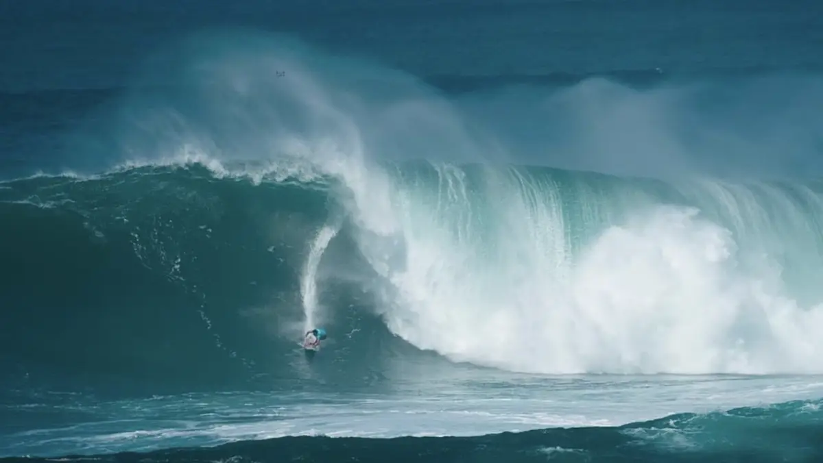 A surfer rides a massive, towering wave in the ocean during one of the North Shore annual events. The wave curls overhead, creating a tunnel of water. The surfer appears small in comparison to the sheer size of the wave, with white foam and spray from the wave's powerful break visible around them.
