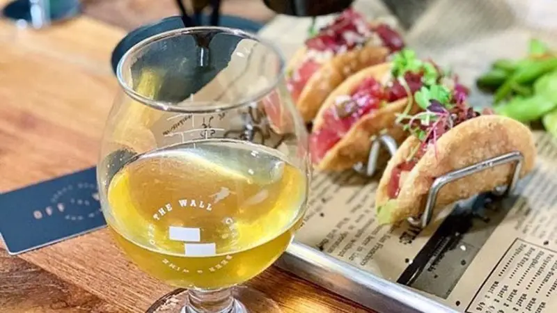 A close-up of a meal on a wooden table, featuring three small tacos filled with vibrant ingredients, placed on a wire holder. A glass of beer with a partially visible logo sits in the foreground. In the background, greenery and dark paper set the scene for the best happy hour Oahu has to offer.