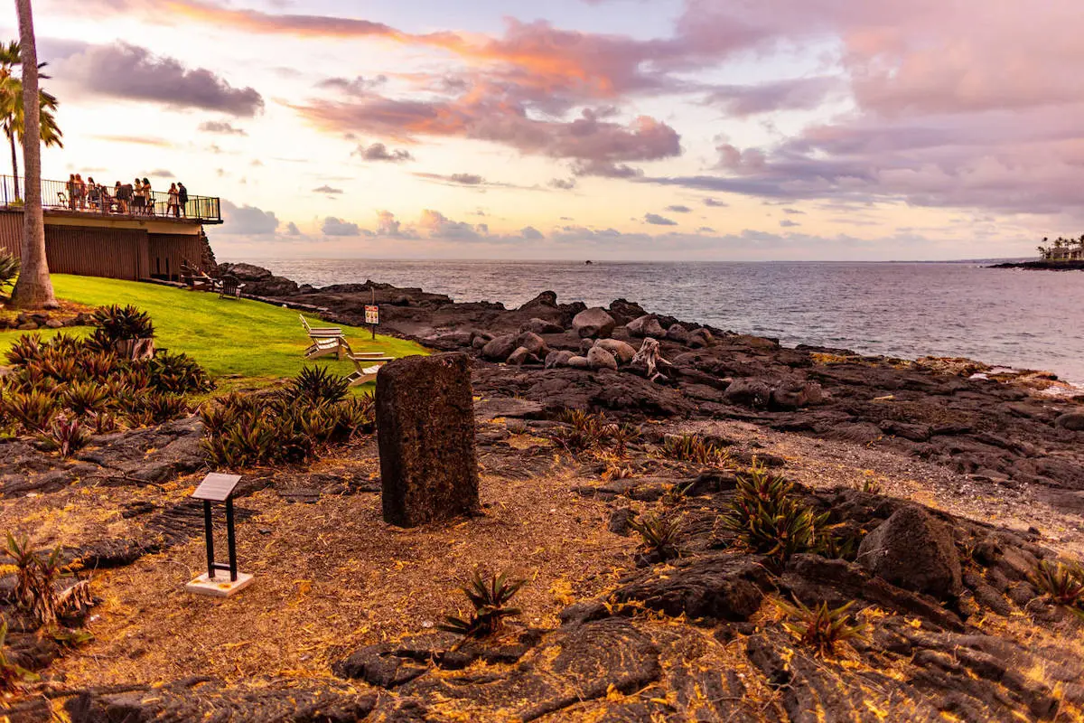 A coastal scene at sunset with a few people standing on a balcony of the Outrigger Hotel Big Island, looking over the ocean. The foreground features a rocky shore with a historical marker and plaque. The sky is a blend of warm colors, creating a serene atmosphere.