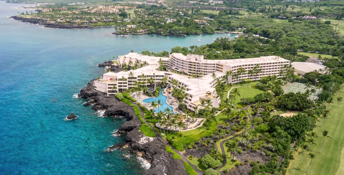 Aerial view of the Kā‘anapali Beach Hotel, a luxury Hawaii home on a beachfront with multiple buildings, a winding pool, lush green vegetation, and a rocky coastline surrounded by turquoise ocean waters. The background features more greenery and additional buildings nestled among the trees.