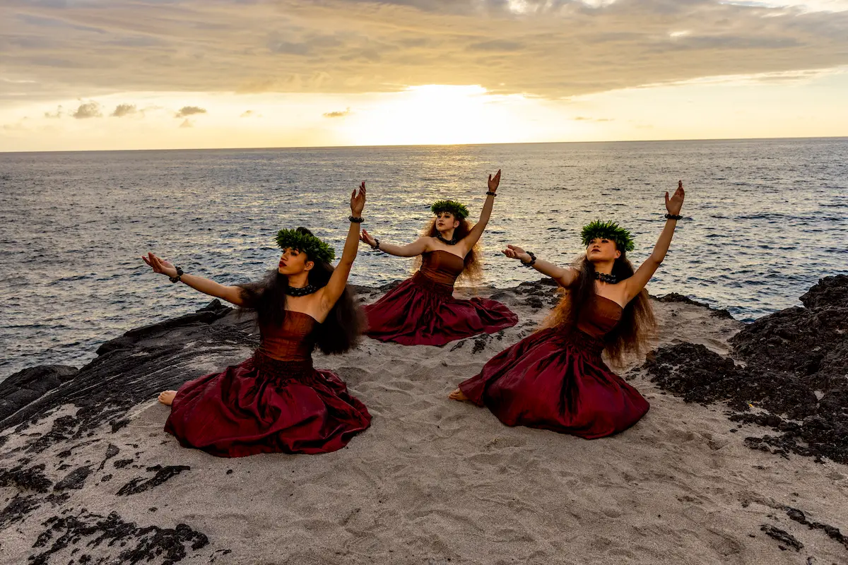 Three individuals dressed in traditional Hawaiian attire perform hula dancing on a sandy beach at sunset, near the Outrigger Hotel Big Island. They are wearing red skirts and leafy headpieces, with the ocean and a dramatic sky in the background. Their arms are elegantly extended.