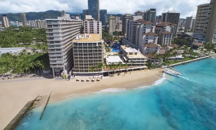 An aerial view of a beachfront cityscape featuring high-rise buildings, resorts, and palm trees along the shoreline showcases some of the best hotels on Oahu. Crystal-clear turquoise waters gently lap the sandy beach, with a few beach chairs set up under umbrellas. A docked boat is visible on the right.