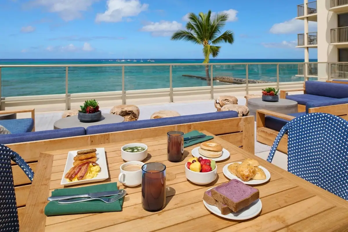 A wooden table on a beachfront patio at the Outrigger Hotel Big Island is set with a variety of breakfast foods, including eggs, bacon, sausages, bread, pastries, fruits, and beverages. Blue cushioned seating surrounds the table. In the background, there's an ocean view with a palm tree and clear sky.