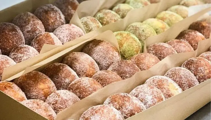 A close-up view of three rows of assorted, sugar-coated doughnuts neatly arranged in a box. The doughnuts vary slightly in color, possibly indicating different flavors. Reminiscent of the best malasadas in Honolulu, they are placed in paper liners within the box.