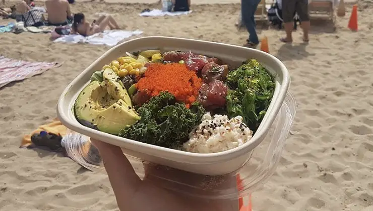 A hand holds up the best poke bowl in Waikiki, filled with avocado, corn, seaweed, raw fish, orange roe, and rice, against the backdrop of a sandy beach on Oahu with people lounging on towels and sunbathing.