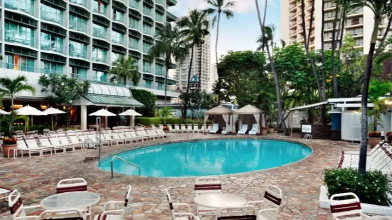 A serene outdoor pool area at the Sheraton Princess Kaiulani hotel is surrounded by lounge chairs, tables, and umbrellas. Tall palm trees and lush greenery provide shade. High-rise buildings loom in the background, indicating an urban setting. The empty pool area creates a tranquil atmosphere.