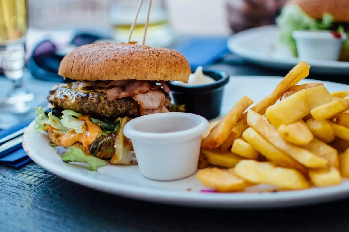 A close-up of a plate containing one of the best burgers in Princeville, a cheeseburger with bacon, lettuce, and tomato, served with golden French fries. Two small dipping cups with condiments accompany the meal, and a glass of drink is partially visible in the background.