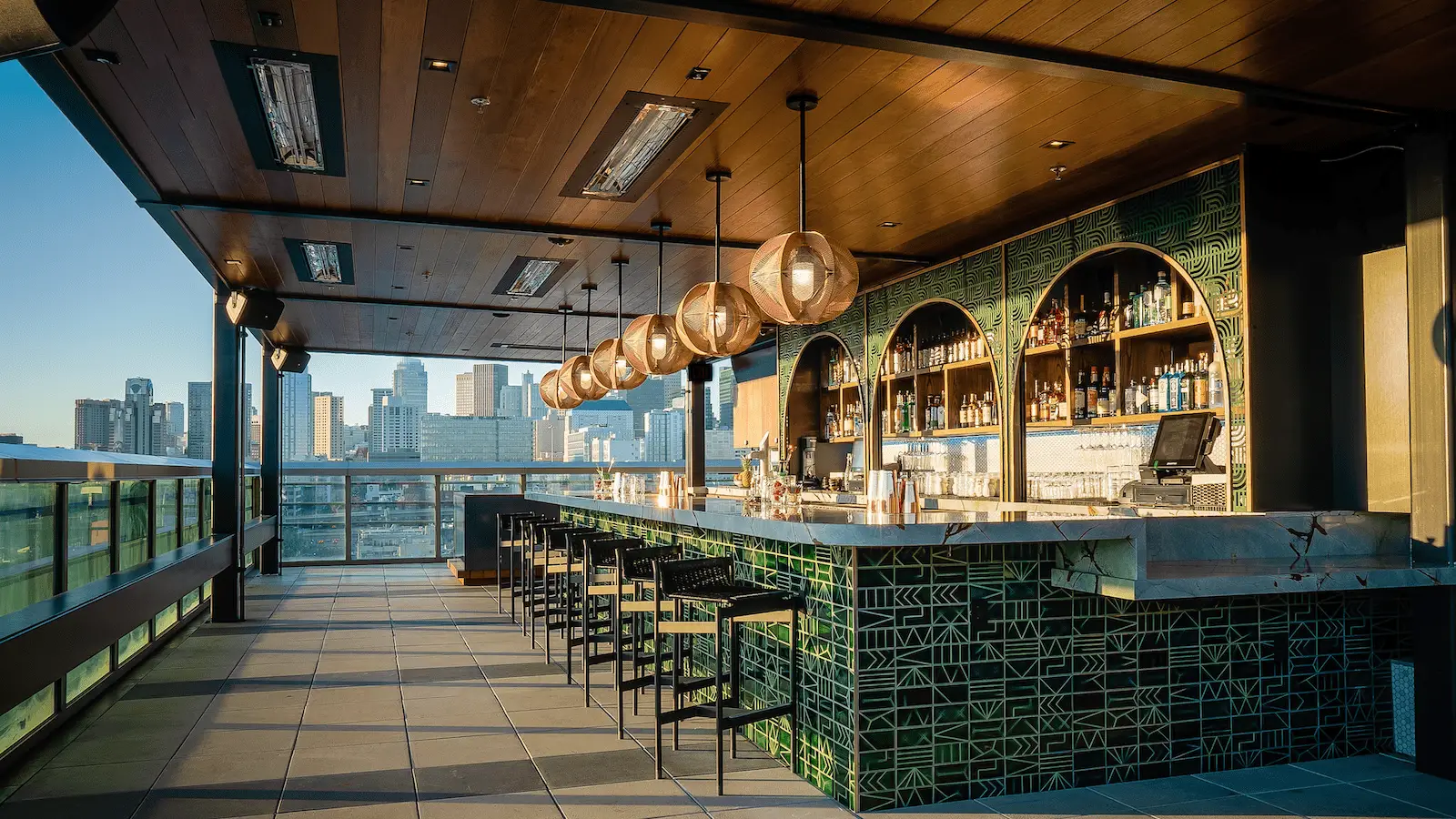 A modern rooftop bar with stylish pendant lights, green geometric tiles, and sleek bar stools. The space offers a panoramic city skyline view during sunset, making it one of the coolest rooftop dining spots in the Bay Area. Enjoy a mix of outdoor seating and a partially sheltered bar area under a wooden ceiling.
