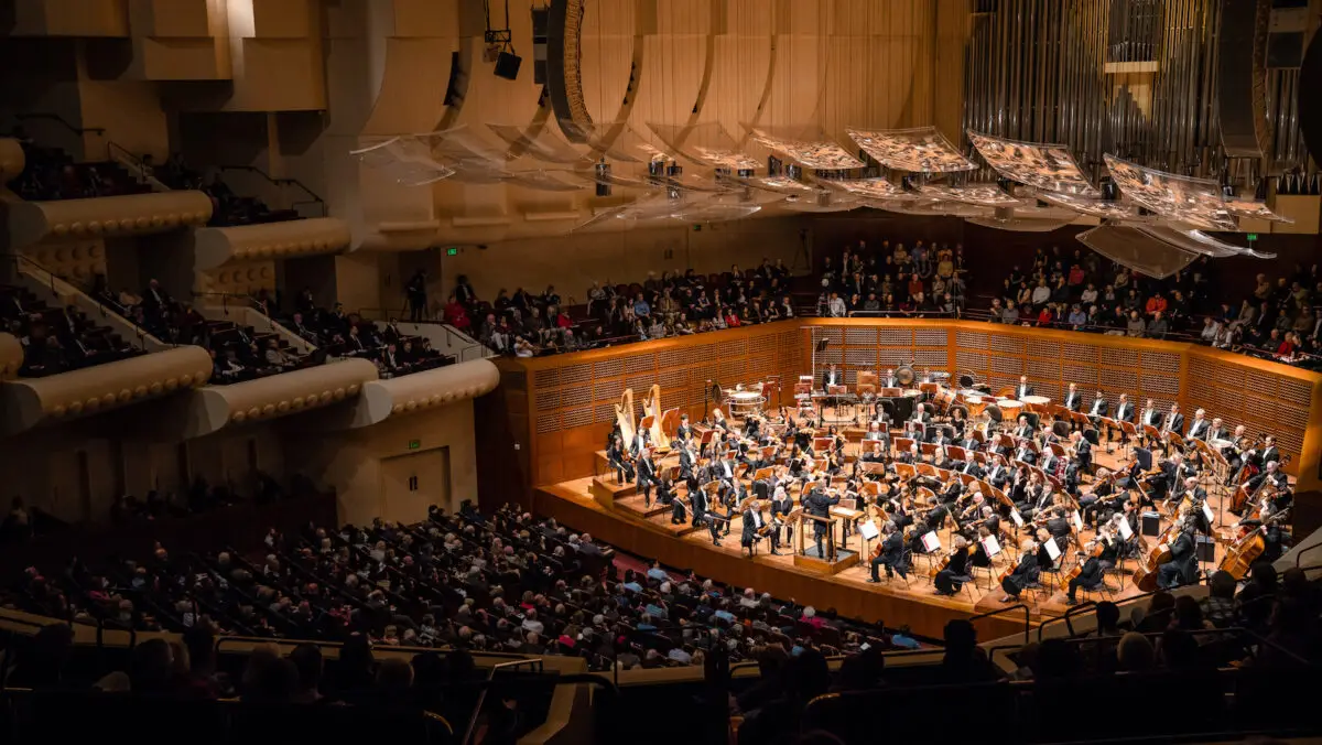 The San Francisco Symphony performs on stage in a spacious, modern concert hall with a high ceiling and unique architectural elements. The audience, seated tightly together, closely watches the performance as the hall's lighting highlights the performers and their instruments.