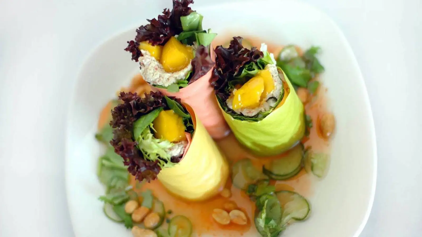 A white plate holds three colorful fresh spring rolls wrapped in green and pink rice paper. The rolls, filled with vegetables, leafy greens, and slices of mango, might just be the Best Dinner on Maui. The plate is garnished with sliced cucumbers, peanuts, and drizzled with a light orange sauce.